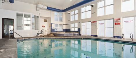 An indoor swimming pool with clear blue water, large windows, and safety signs.