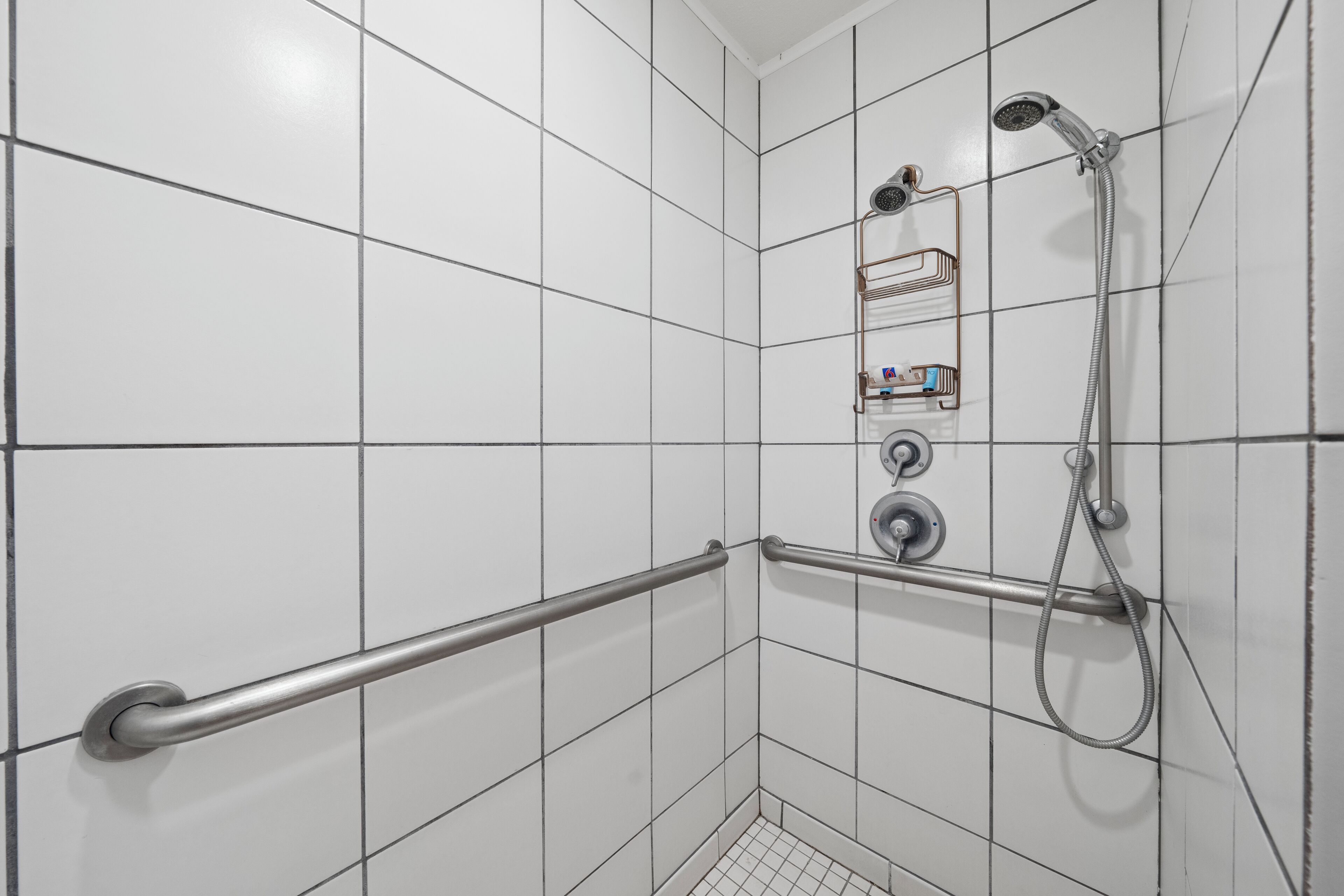 Un baño con paredes de azulejos blancos, una ducha con cabezal de ducha manual, otro cabezal de ducha manual y un perchero para toallas.