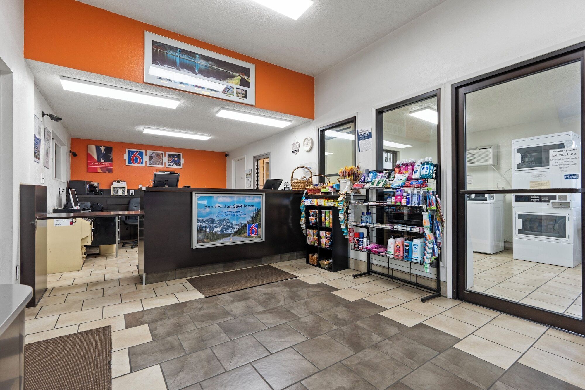A hotel reception area with a check-in counter, a digital display, and a self-service kiosk. There are various amenities including a coffee machine, a refrigerator, and a vending machine.