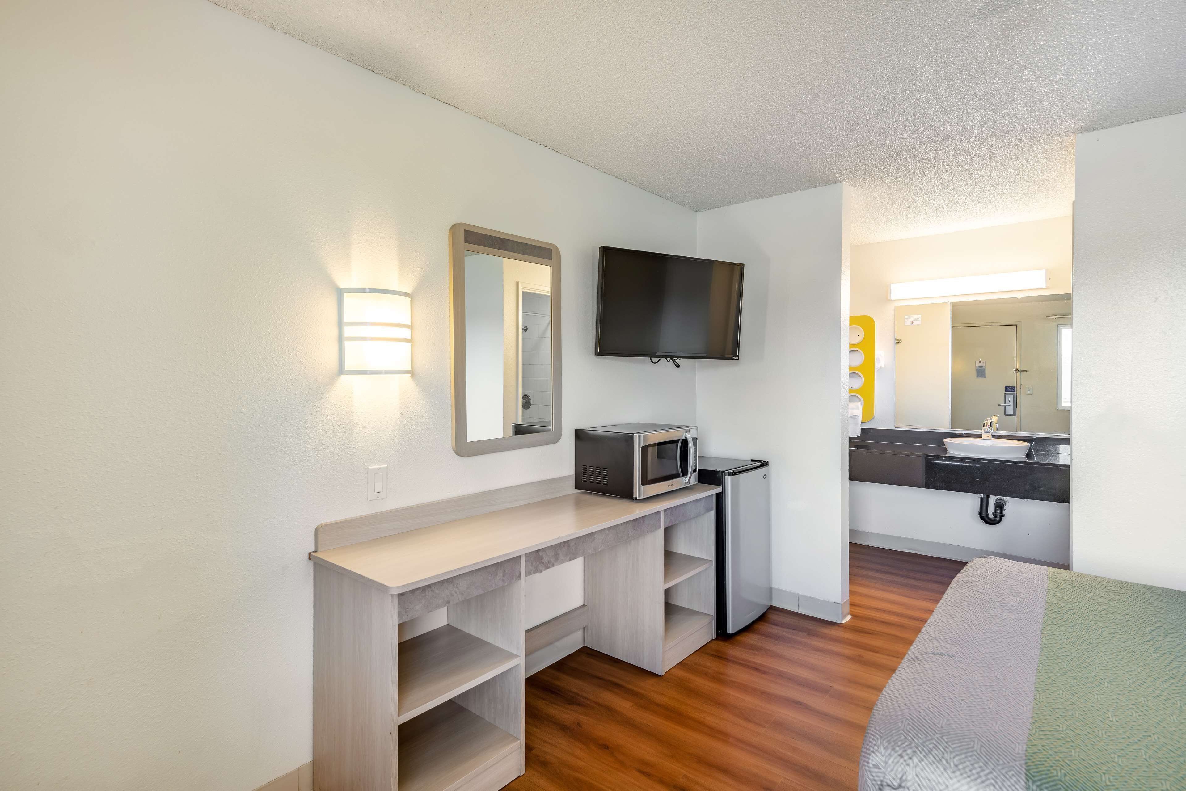 A compact hotel room with a kitchenette, a flat-screen TV, a mirror, and a bed.