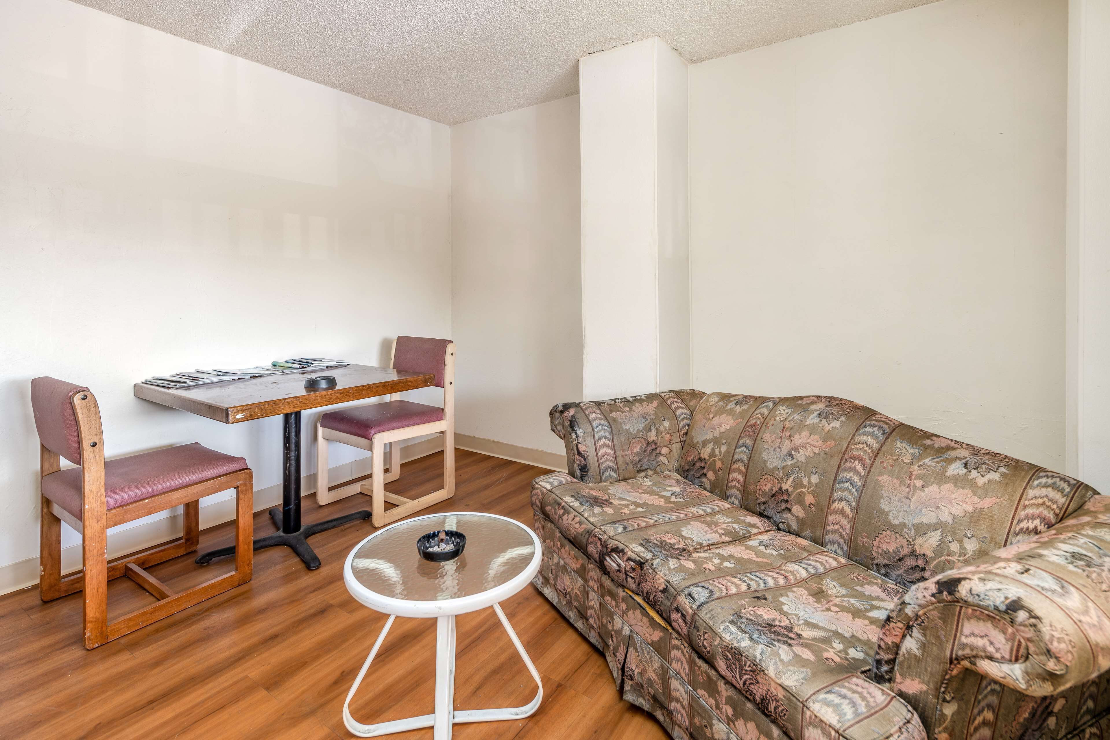A small, furnished room with a wooden floor, a floral sofa, a wooden dining table with two chairs, and a small white table with a bowl.