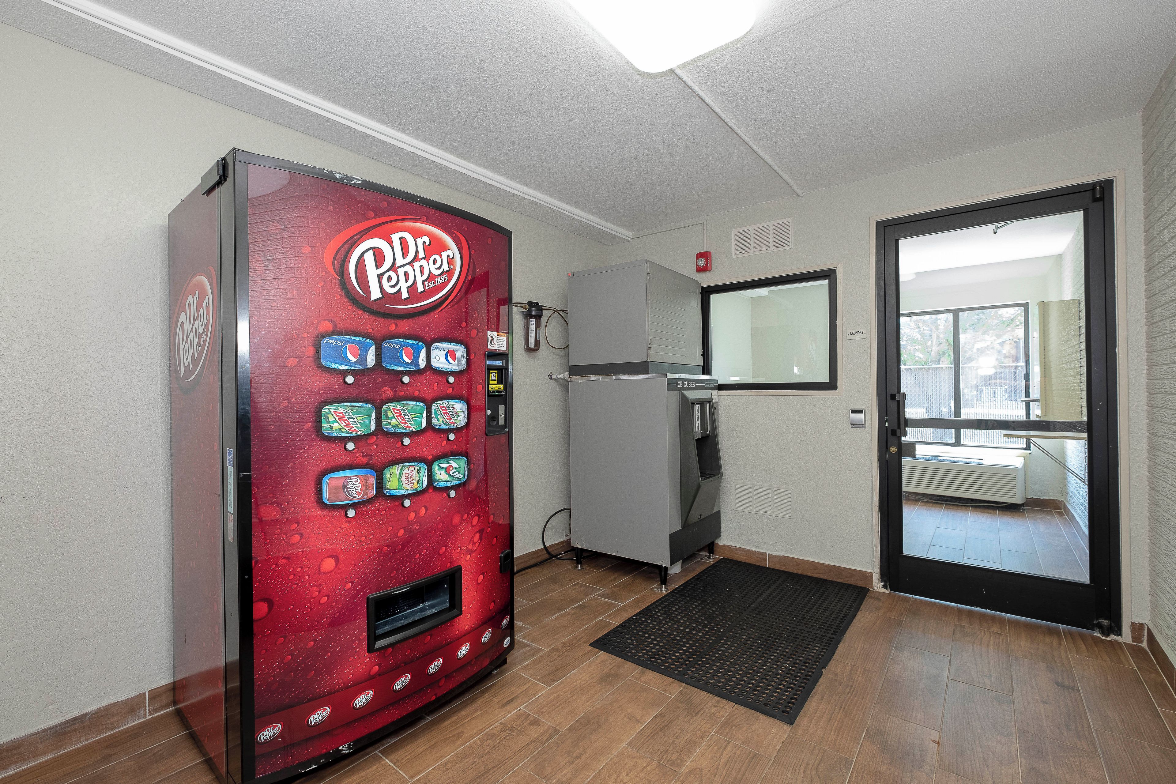 Una máquina expendedora con diferentes bebidas, un refrigerador, una puerta con espejo y un tapete negro en la entrada.