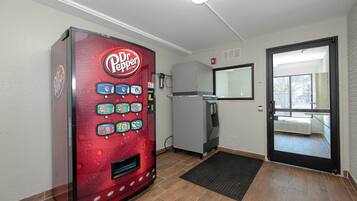 Una máquina expendedora con diferentes bebidas, un refrigerador, una puerta con espejo y un tapete negro en la entrada.