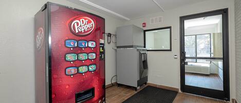 Una máquina expendedora con diferentes bebidas, un refrigerador, una puerta con espejo y un tapete negro en la entrada.