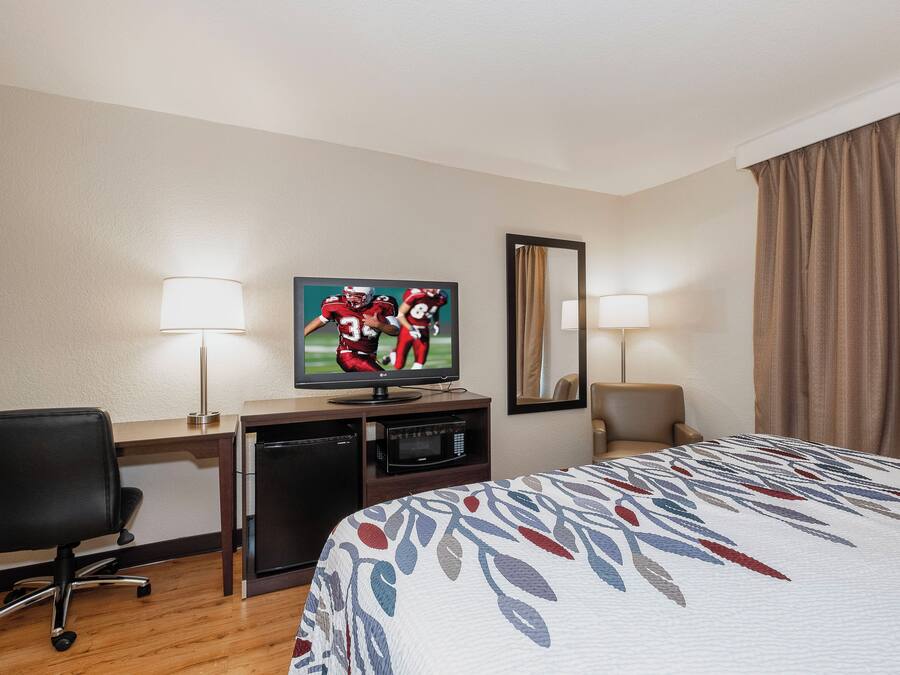 A hotel room with a bed, a desk with a TV, a chair, a desk lamp, a mirror, and a window with curtains.