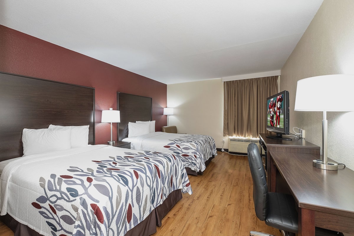 A hotel room with two beds, a desk, a chair, a TV, and a lamp.