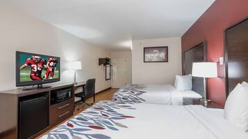 A hotel room with a flat-screen TV, a desk with a chair, a bed with white bedding, and a patterned carpet.