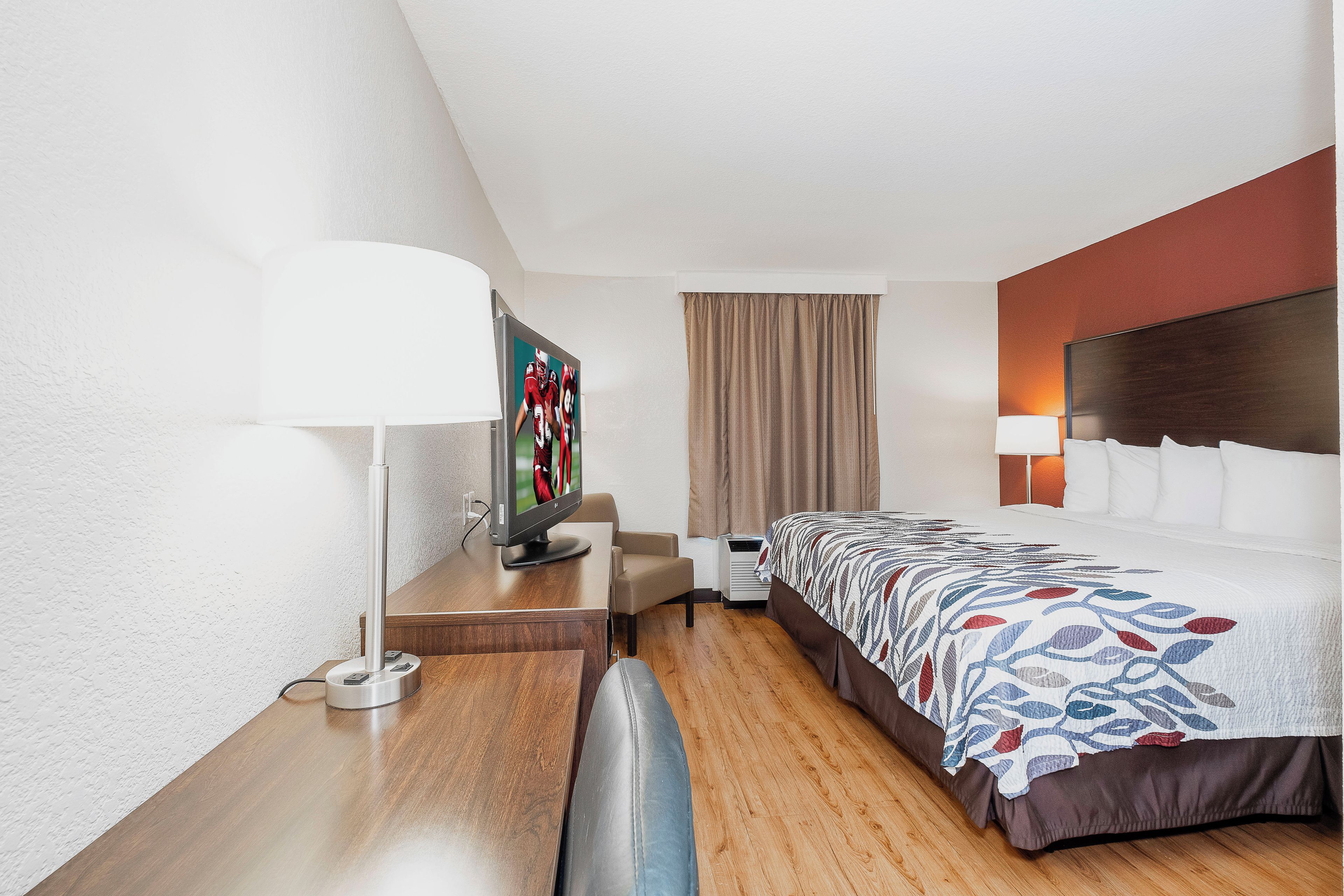 Una habitación de hotel con cama, escritorio, televisor, lámpara y ventana con cortinas.