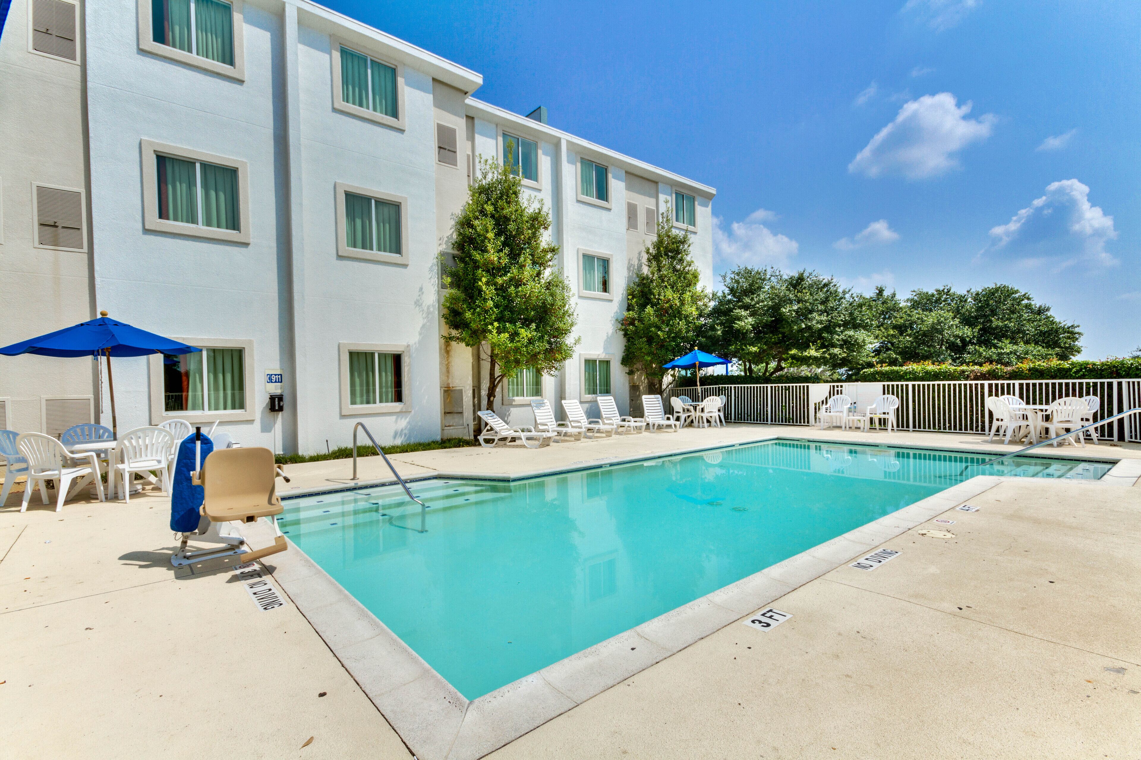 A swimming pool with lounge chairs and umbrellas, surrounded by a building with multiple windows and a clear sky.