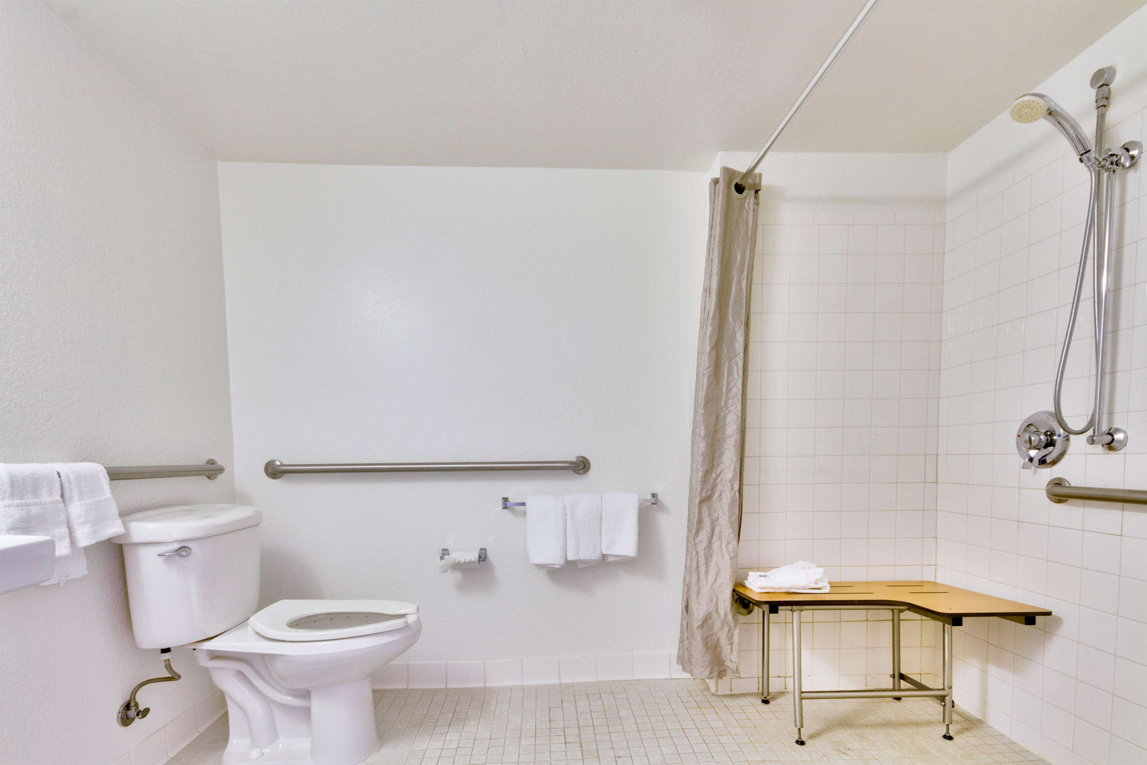 A bathroom with a toilet, a shower with a handheld showerhead, a grab bar, and a small table with a towel.