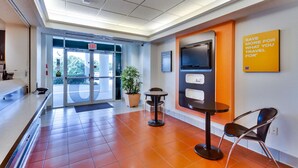 A hotel lobby with a reception desk, a self-service kiosk, a potted plant, and a sign encouraging saving for travel.