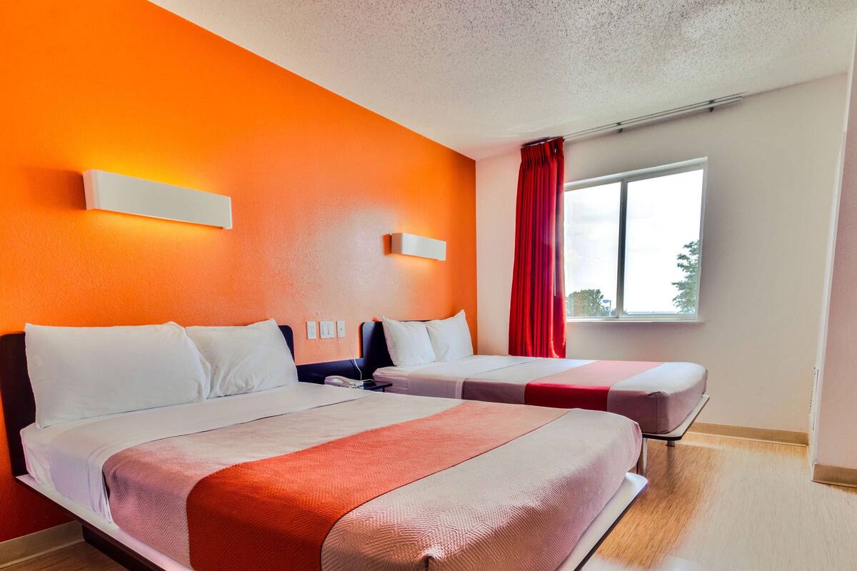 Una camera d'albergo con due letti, una finestra con tende rosse e una parete di colore arancione acceso.