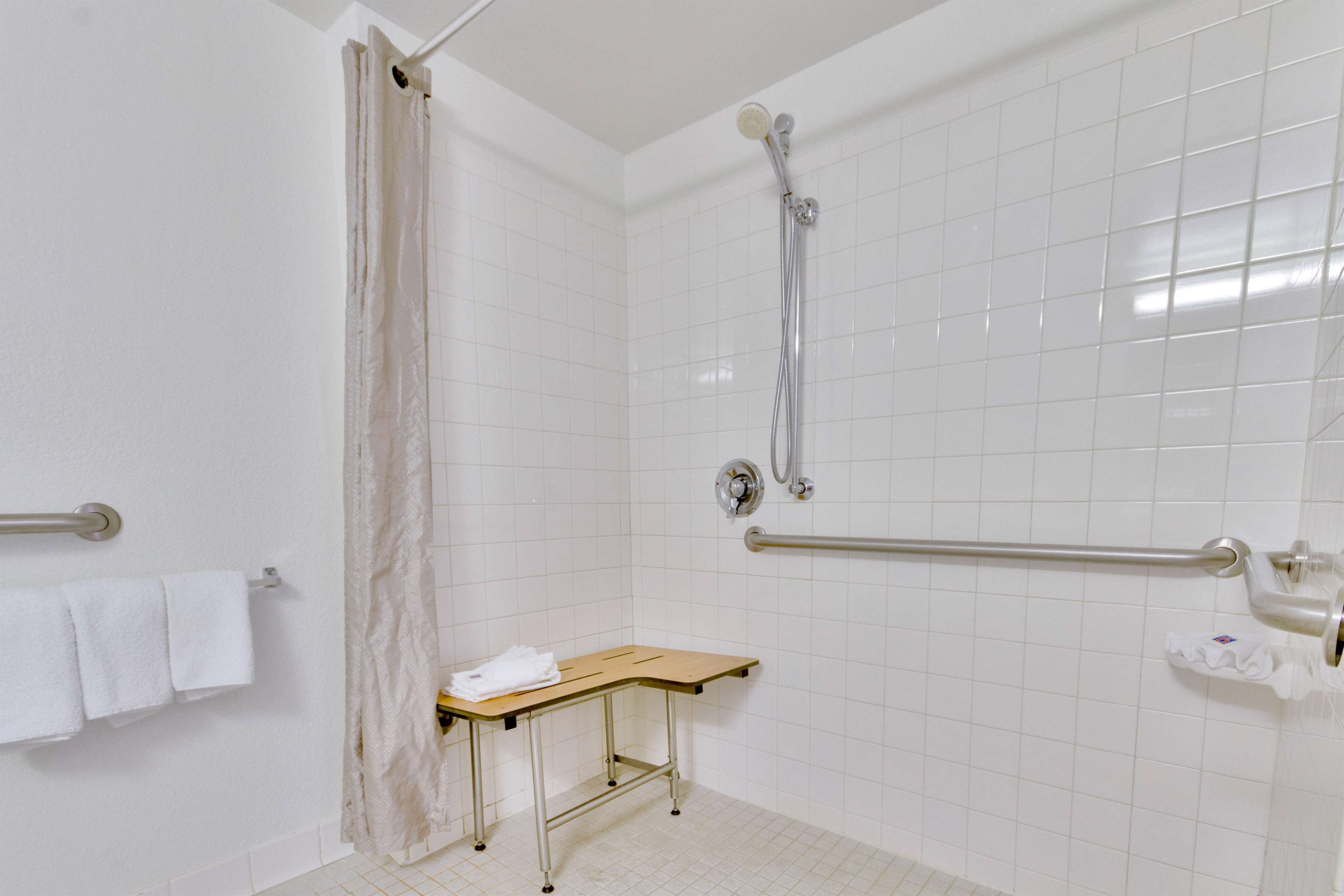 Ein Badezimmer mit weißen Fliesen, einer Dusche inklusive Handbrause, einer Haltestange, einer hölzernen Sitzbank und einem Handtuchhalter mit Handtüchern.