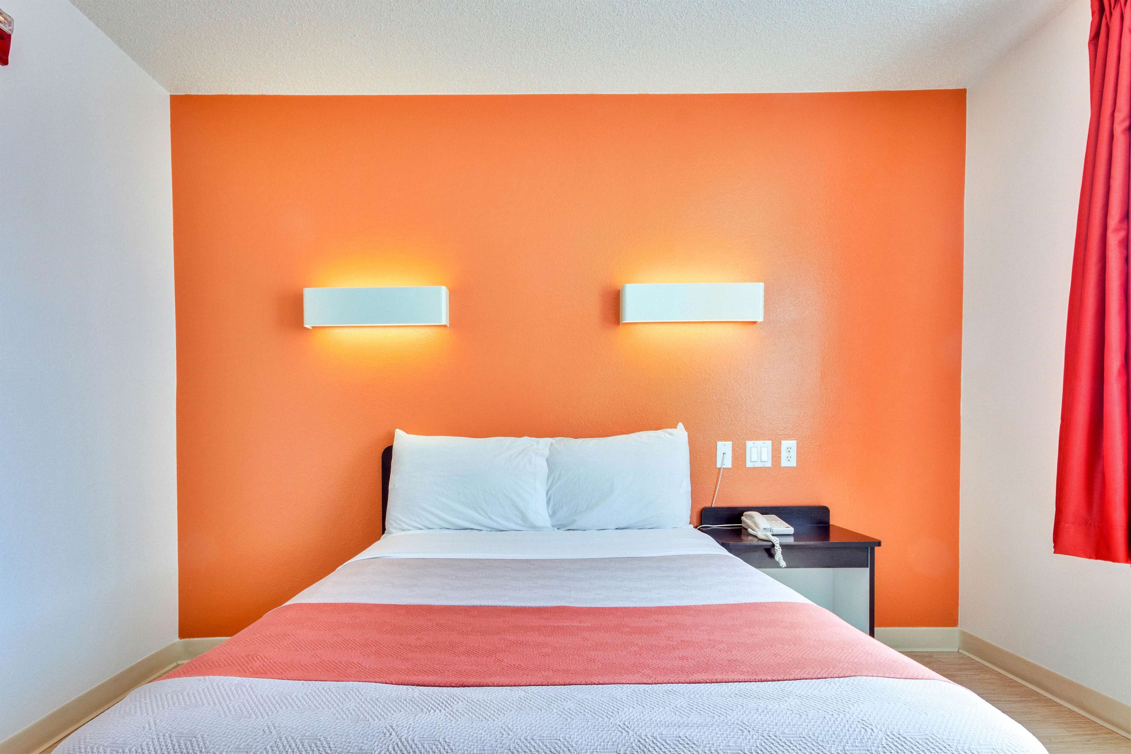 A hotel room with an orange accent wall, a bed with white linens, a red throw blanket, and a nightstand with a phone.