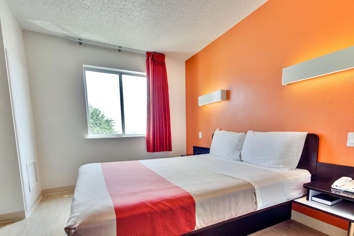 Una camera d'albergo con un letto, un comodino, un telefono e una finestra con tenda rossa.