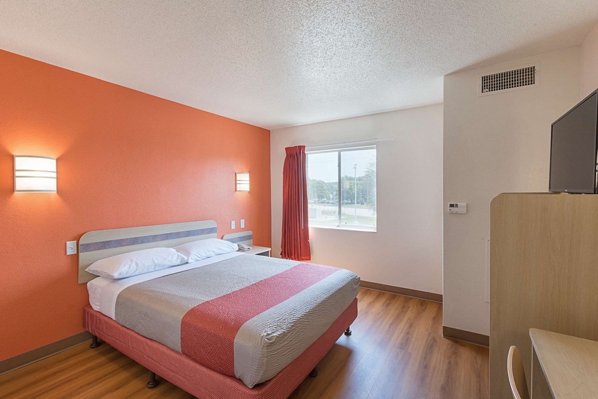A hotel room with a bed, a window with curtains, a nightstand, and a desk.