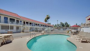 A swimming pool with lounge chairs and tables, a fenced deck area, and a building with a red roof in the background. - Motel 6 Tempe, AZ - Broadway - ASU (Tempe)