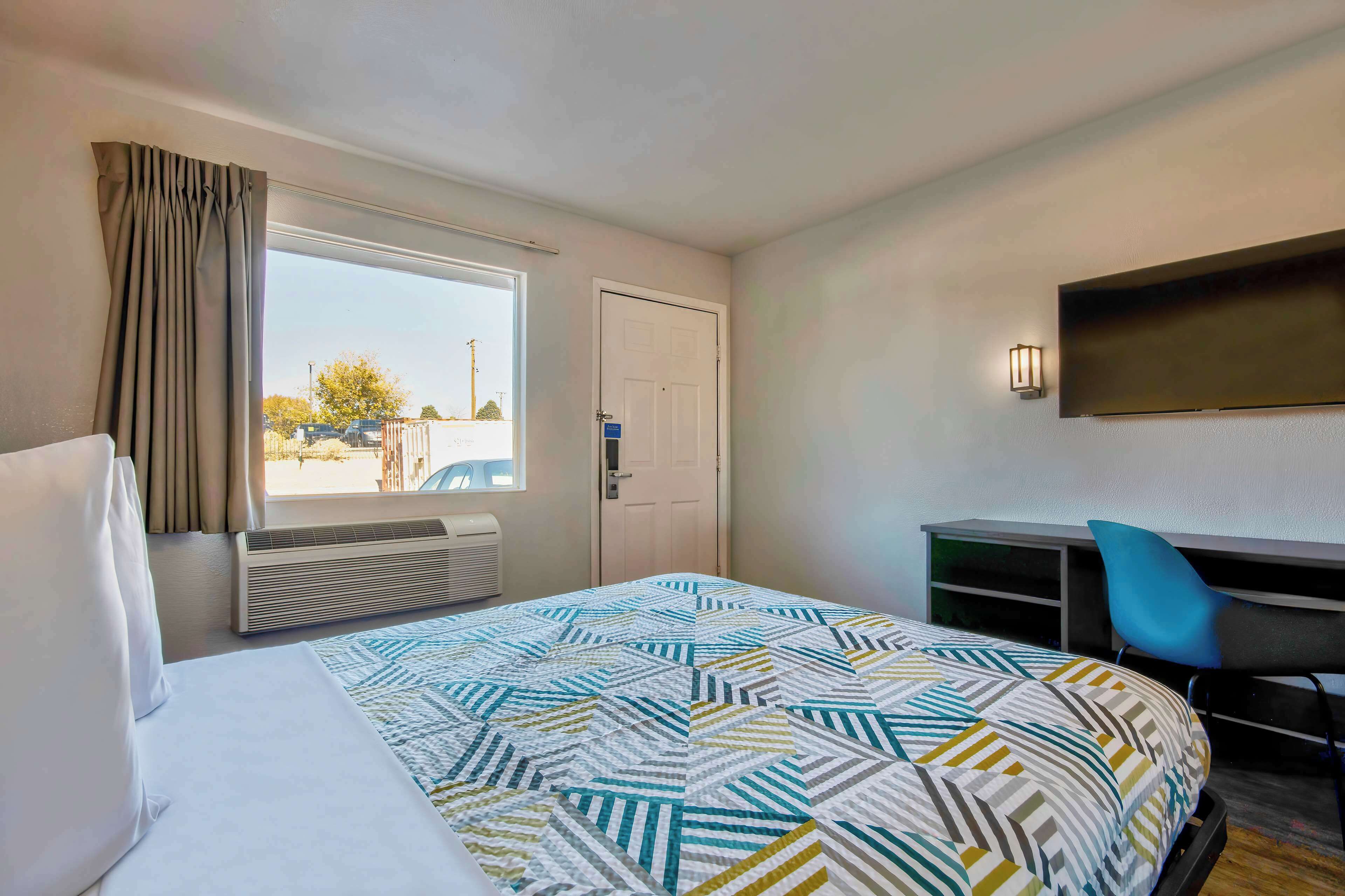 A hotel room with a bed, a desk, a chair, a window with curtains, and a wall-mounted TV.