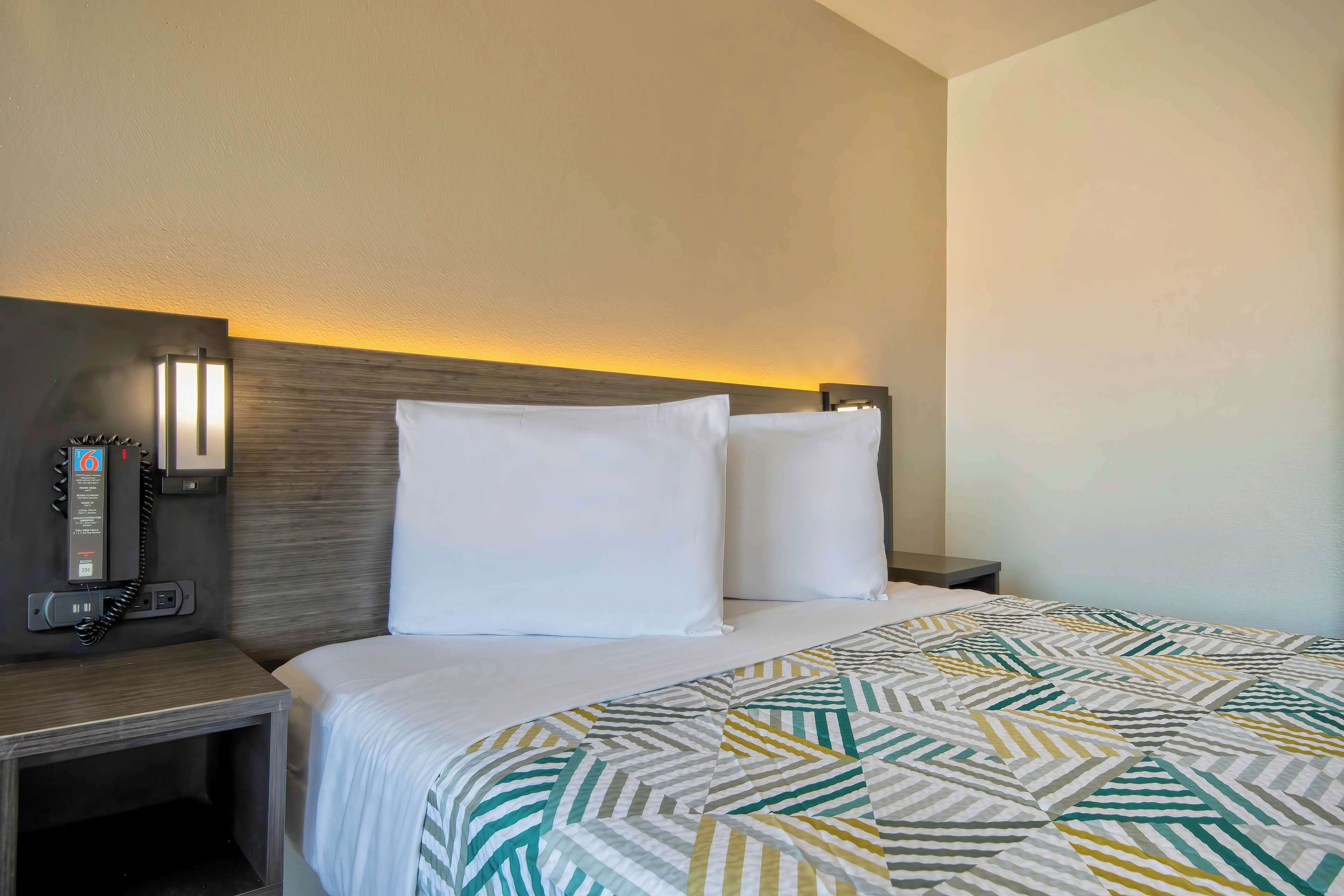 A hotel room with a neatly made bed, a bedside table with a phone, and a wall-mounted light fixture.