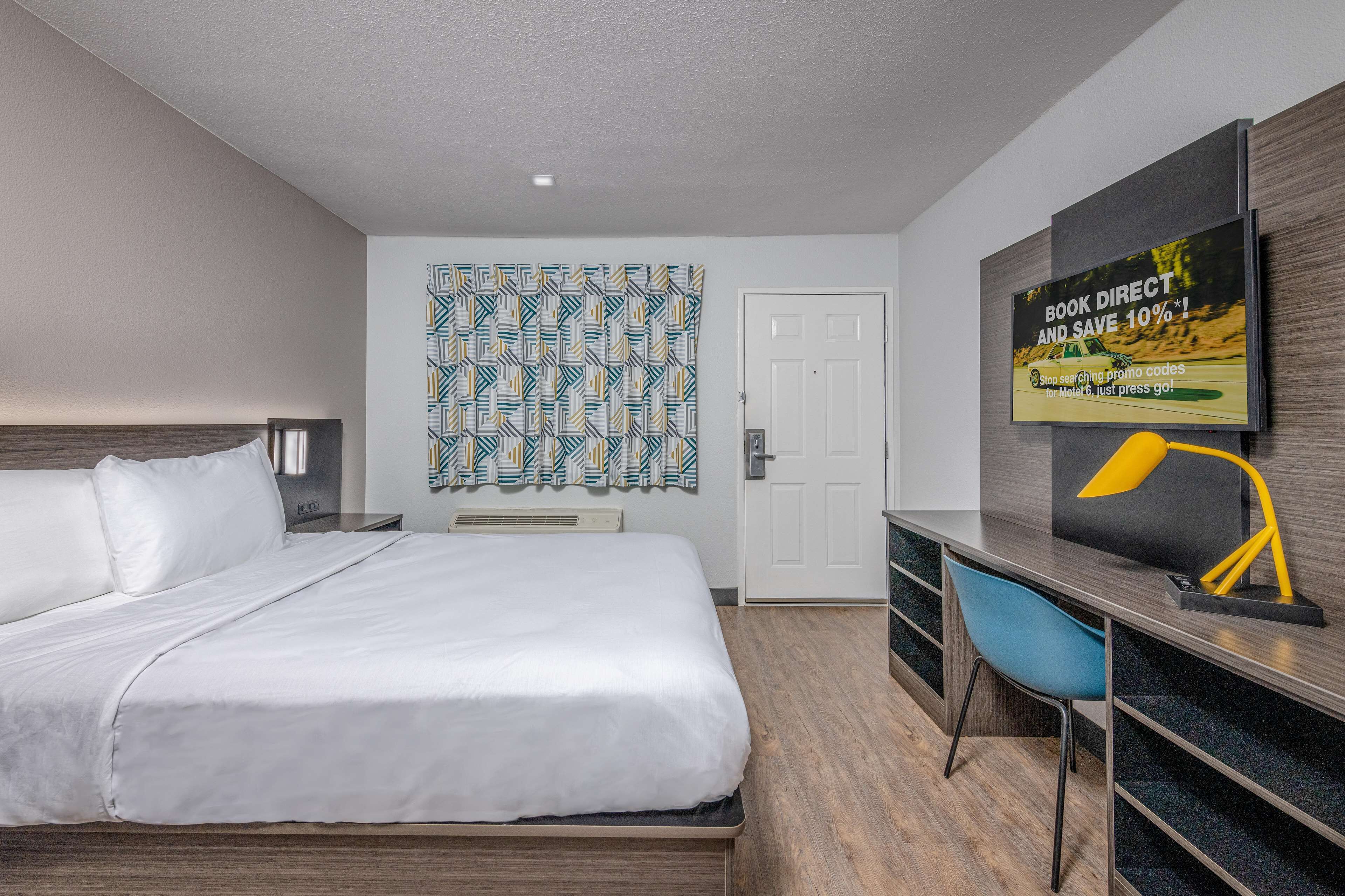 A modern hotel room with a bed, a desk, a chair, a TV, and a patterned wall.