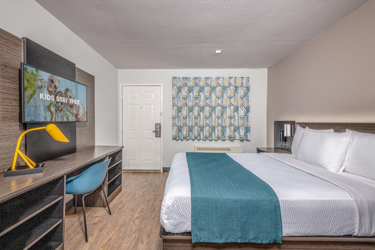 A hotel room with a bed, a desk with a chair, a television, and a patterned wall hanging.