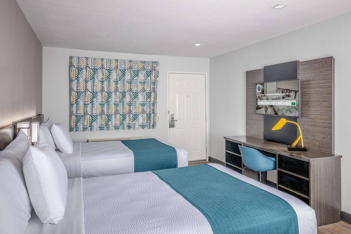 A hotel room with two beds, a desk with a chair, a large mirror, and a patterned curtain.