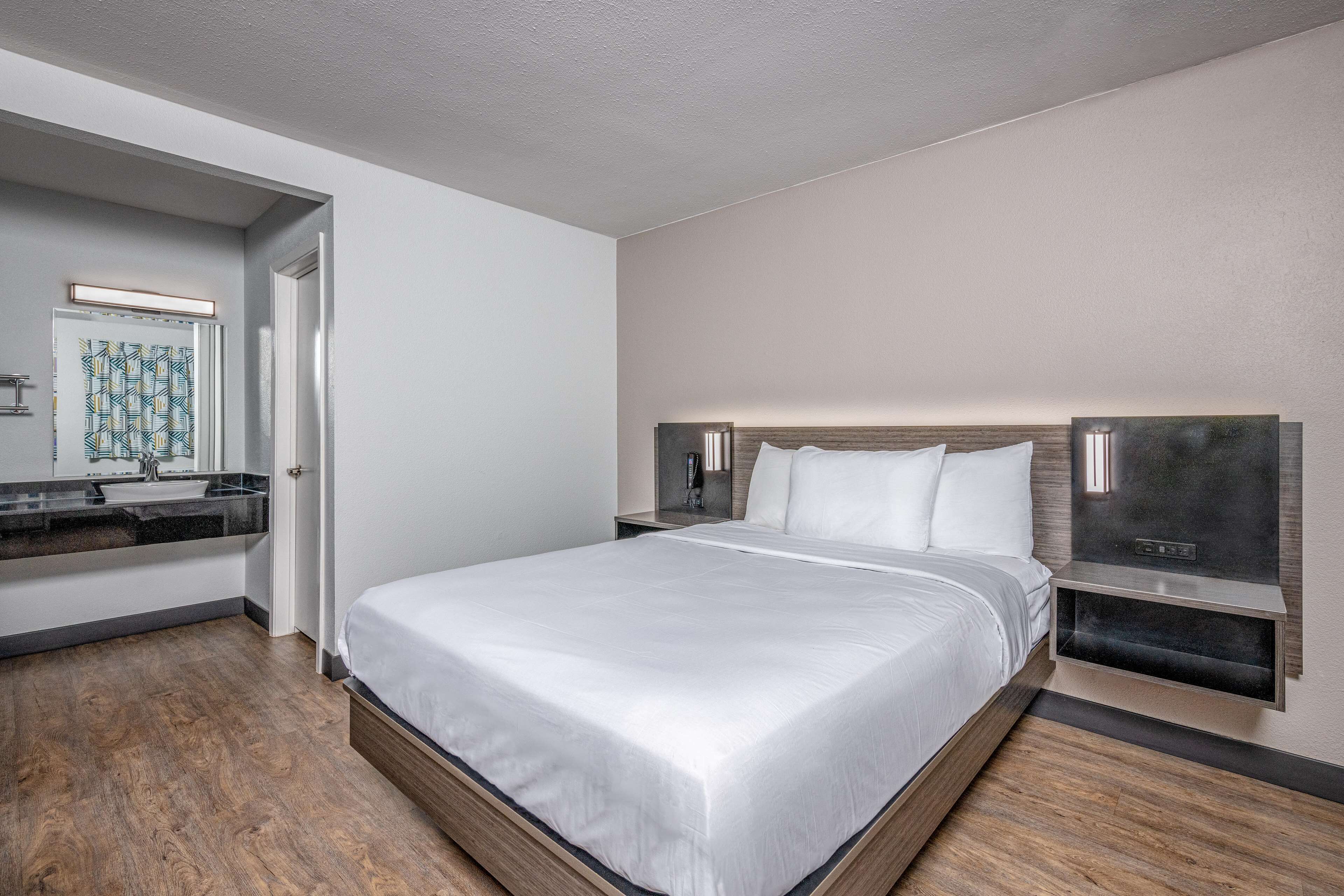 Quarto de hotel moderno com uma cama grande, piso de madeira, banheiro com espelho e pia, e mesa de cabeceira com abajur.