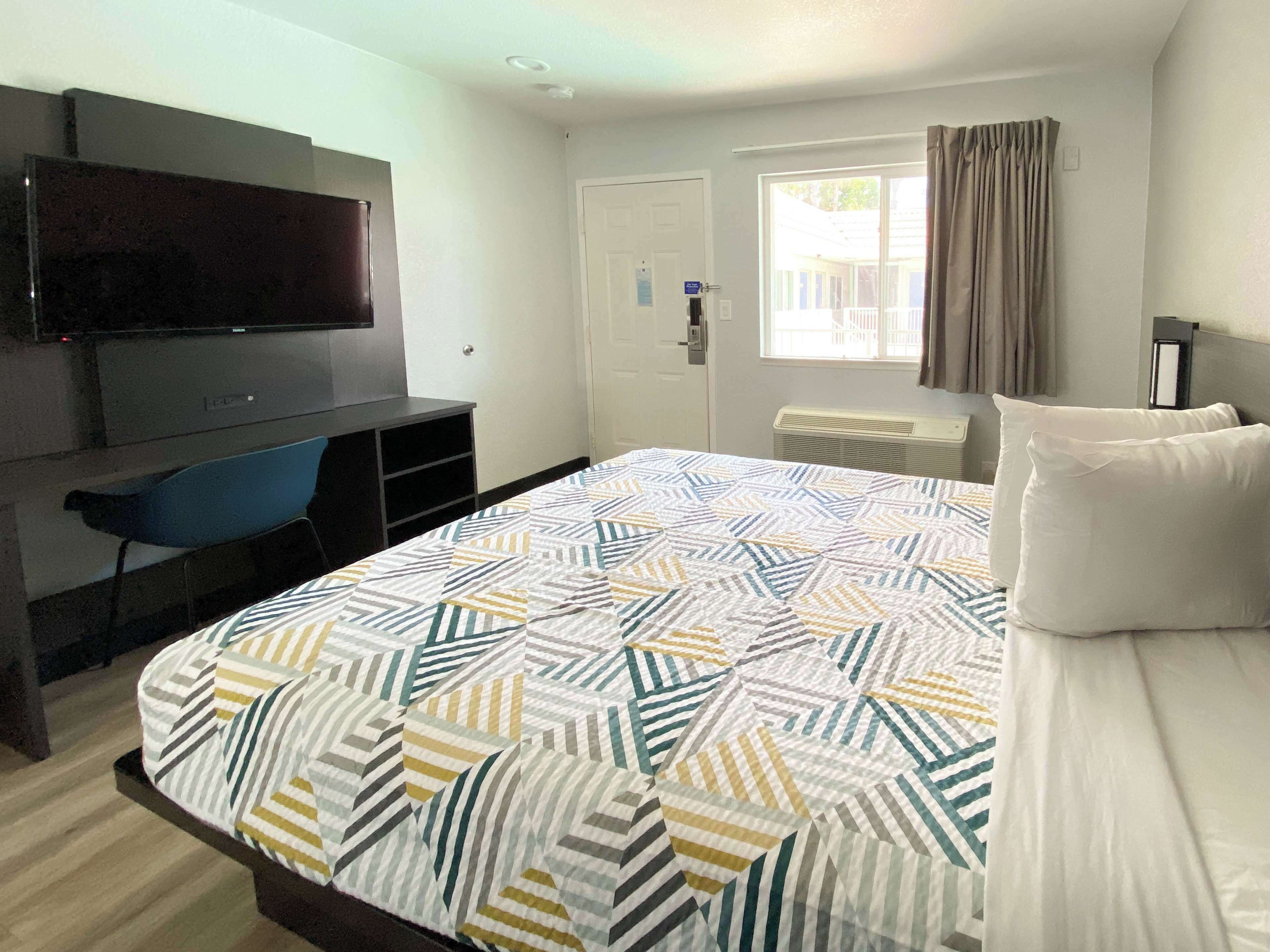 A hotel room with a bed, a TV mounted on the wall, a desk with a chair, and a window with curtains.