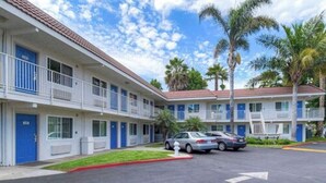Un motel con edificios de dos pisos, cada uno con un balcón y una puerta azul. Hay dos coches estacionados al frente.