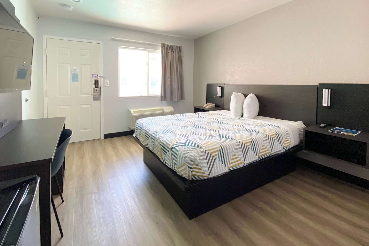 Quarto de hotel com cama, escrivaninha, cadeira, janela com cortinas, mesa de cabeceira com abajur e televisão na parede.