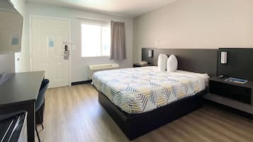 A hotel room with a bed, a desk, a chair, a window with curtains, a bedside table with a lamp, and a wall-mounted TV.