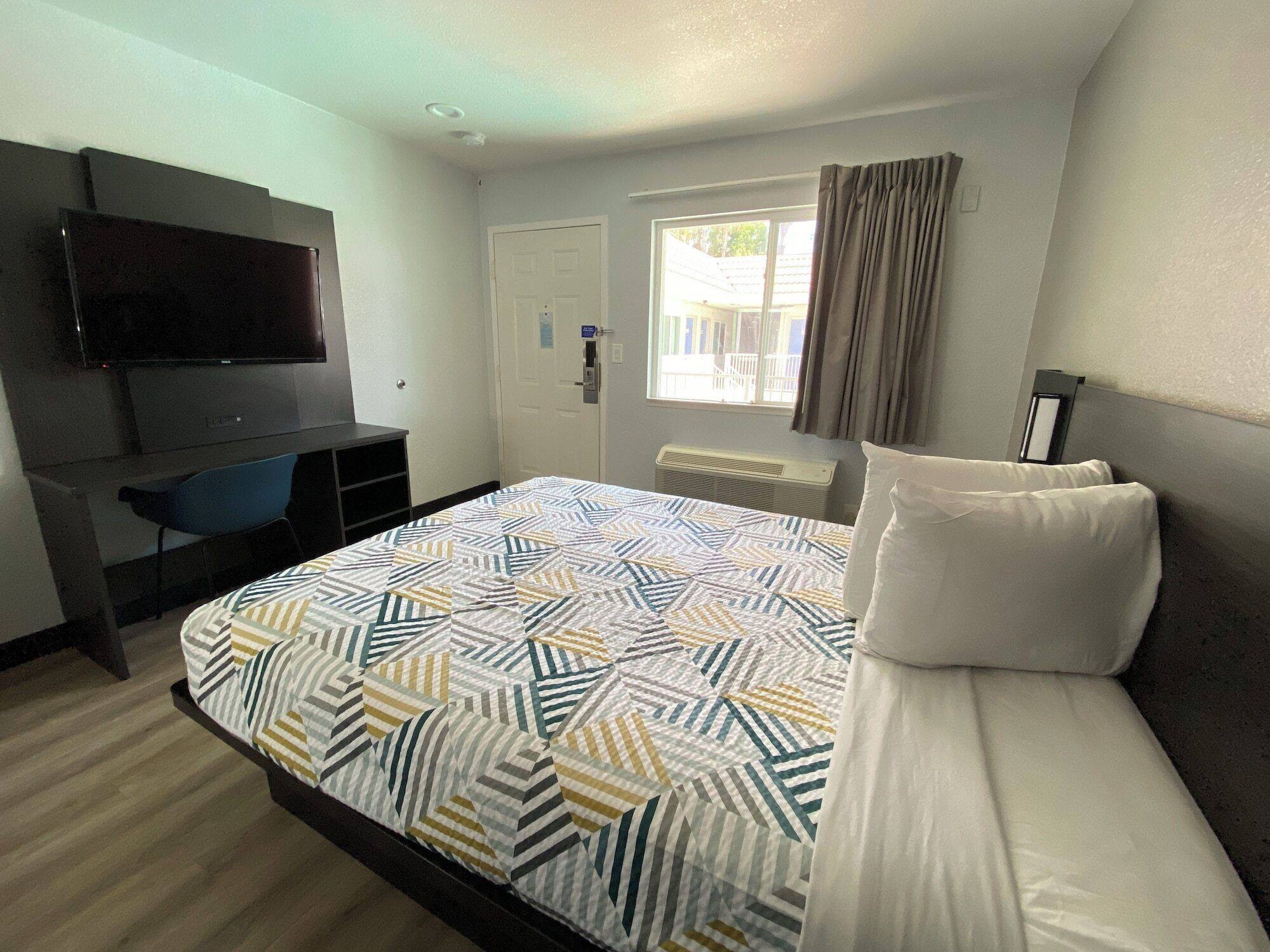 A hotel room with a bed, a desk with a TV, a window with curtains, and a door.