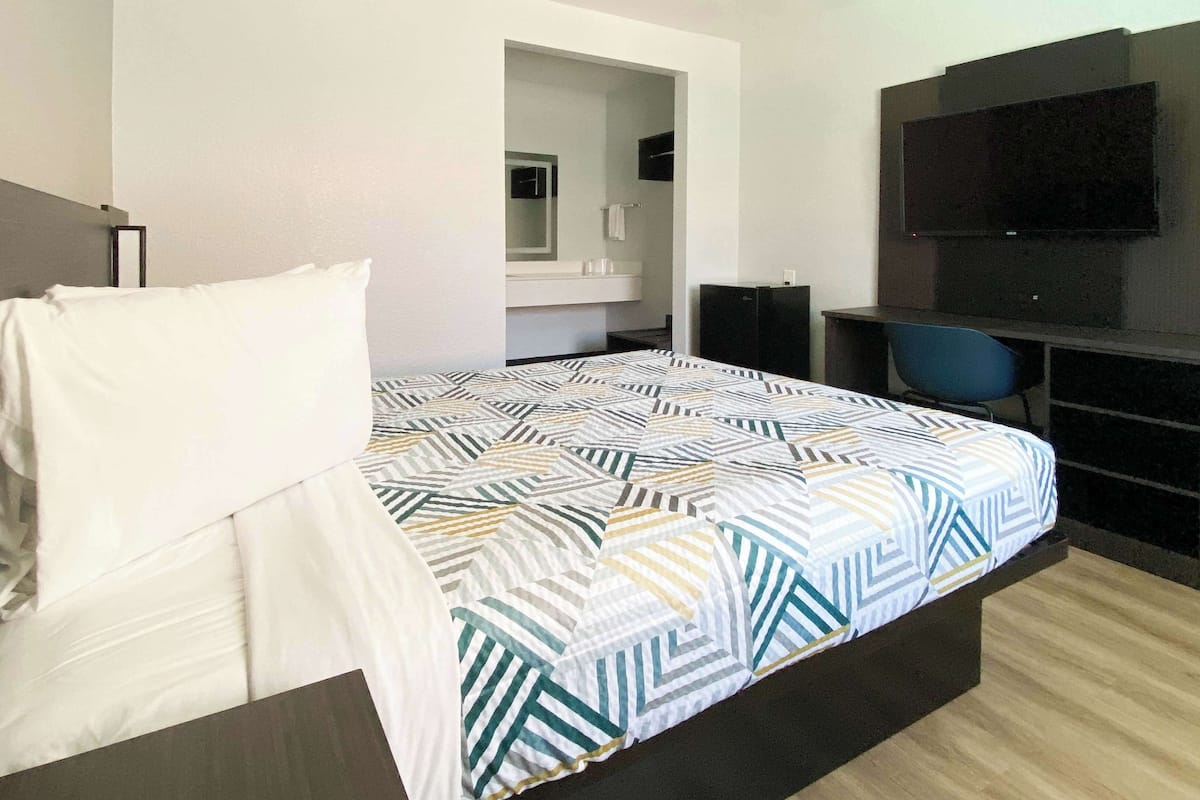 Quarto de hotel com uma cama, escrivaninha, cadeira, televisão e banheiro visível por uma porta aberta.