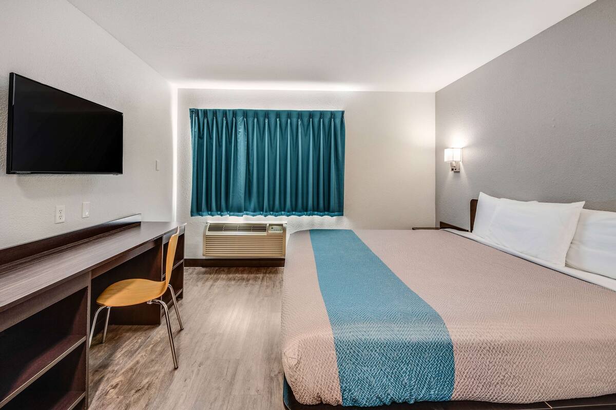 A hotel room with a bed, a desk with a chair, a flat-screen TV mounted on the wall, an air conditioning unit, and a window with teal curtains.