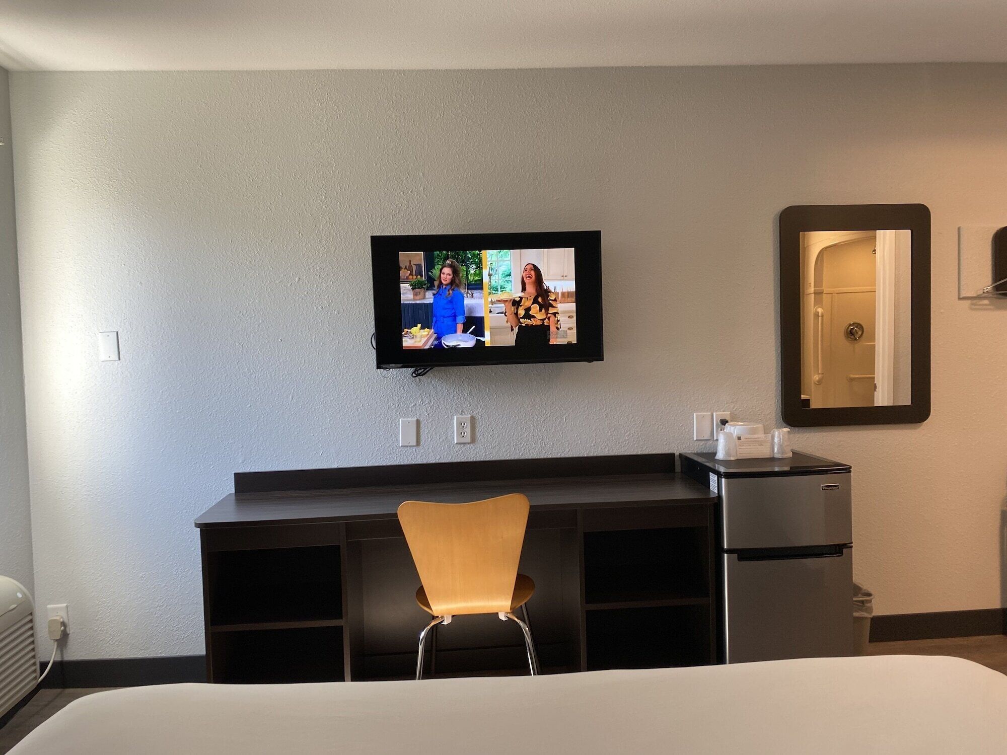 Hotelkamer met een bed, een bureau met stoel, een aan de muur bevestigde televisie, een spiegel en een klein tafeltje met een kopje en zakdoekjes.