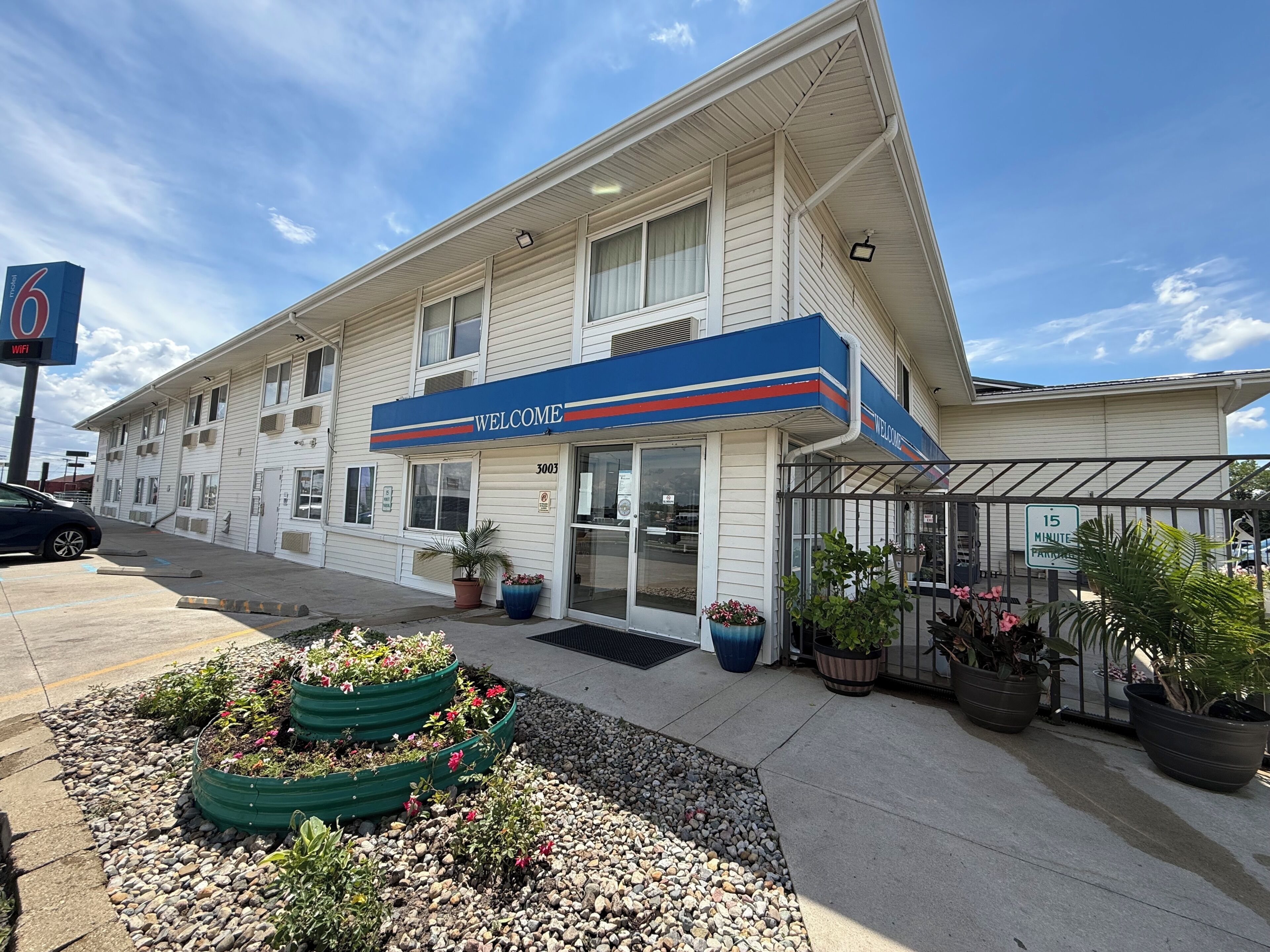 Un motel avec une enseigne « BIENVENUE », un parking et un espace jardin agrémenté de plantes en pot.