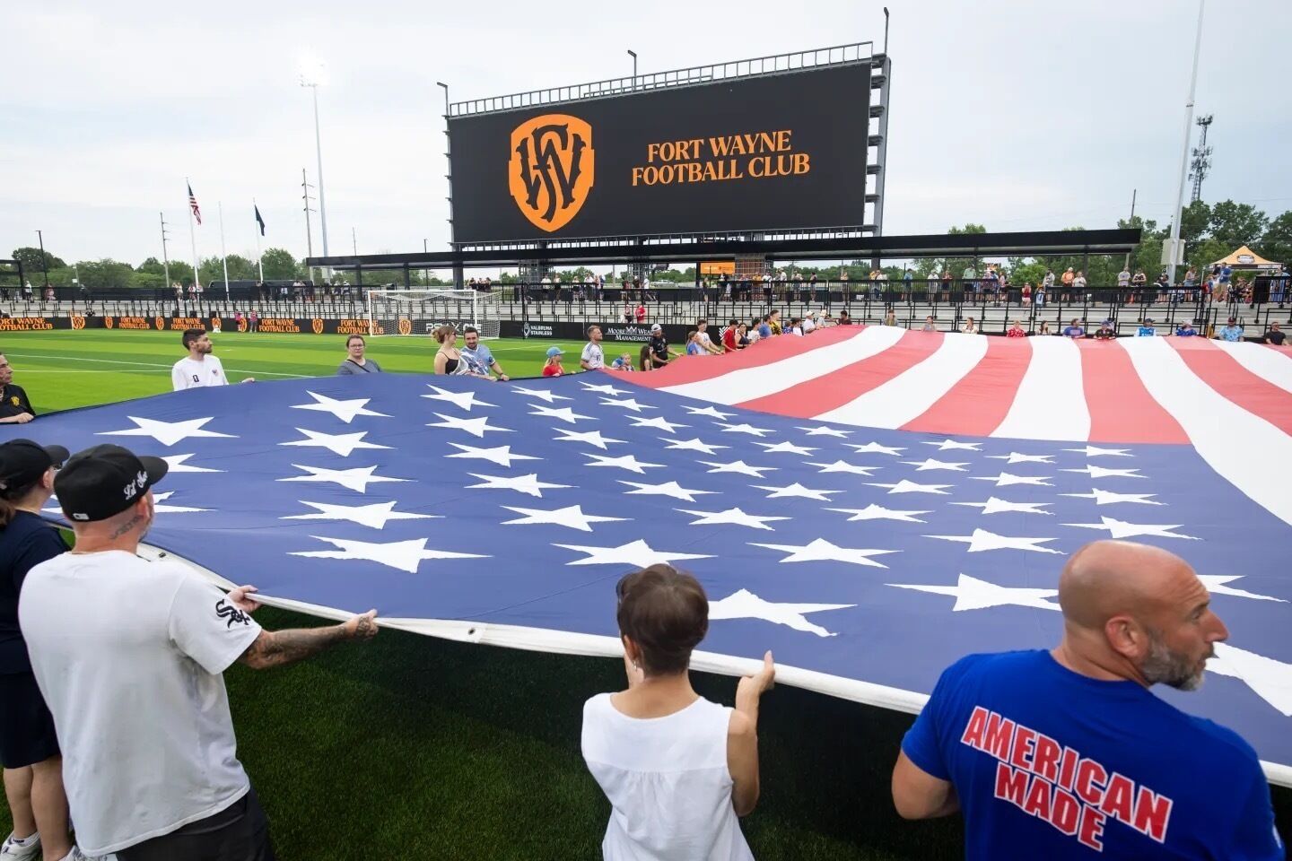 A large American flag being unfurled on a sports field with a scoreboard in the background.