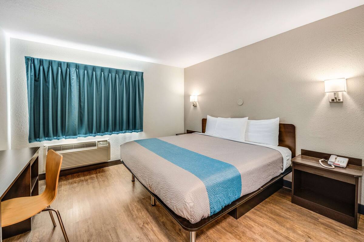 A hotel room with a bed, a desk, a chair, a window with curtains, a wall-mounted phone, and a heater.