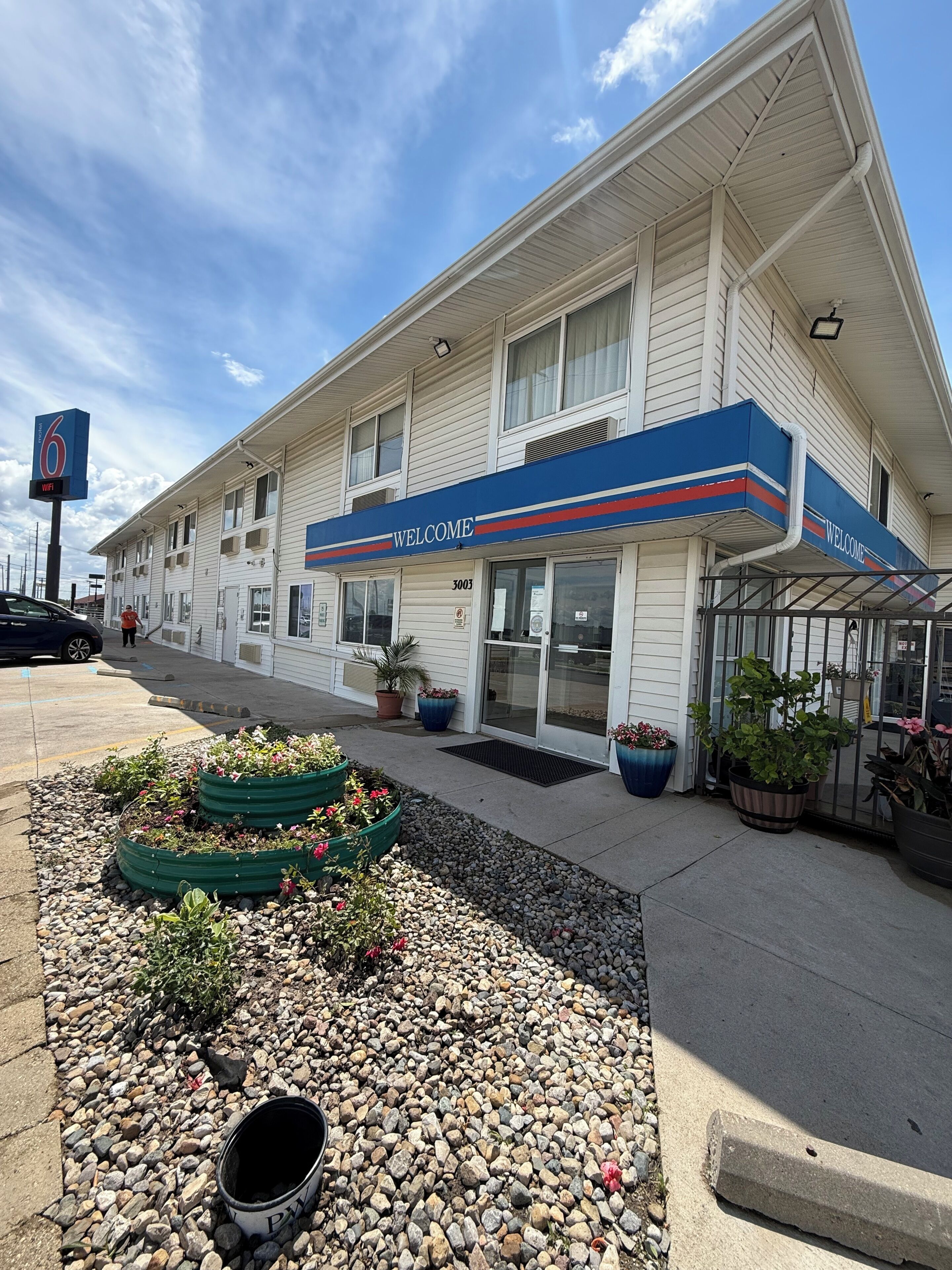 A motel with a 'WELCOME' sign, a parking lot, and potted plants.