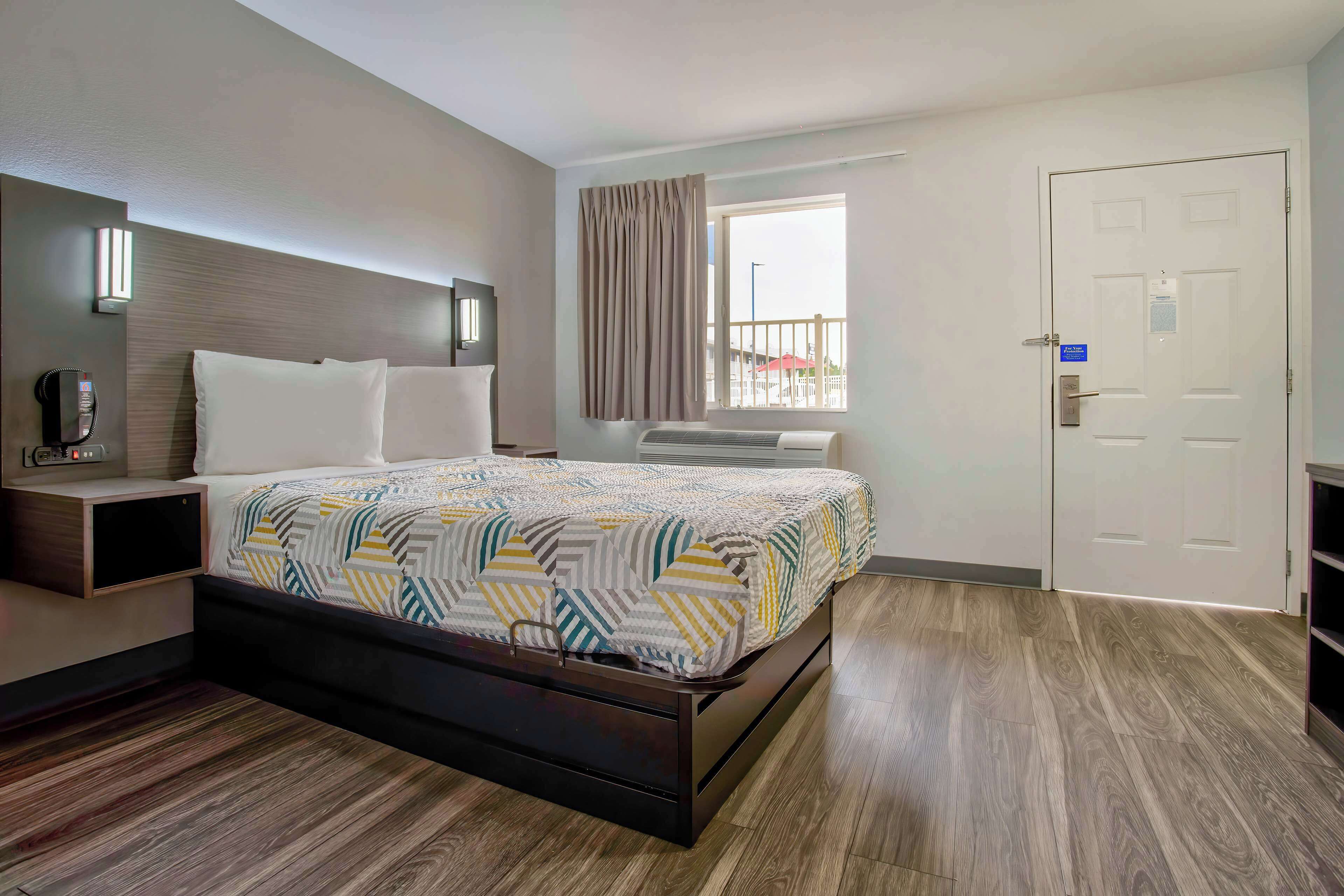 A modern hotel room with a large bed, bedside tables, a window with curtains, and a door.