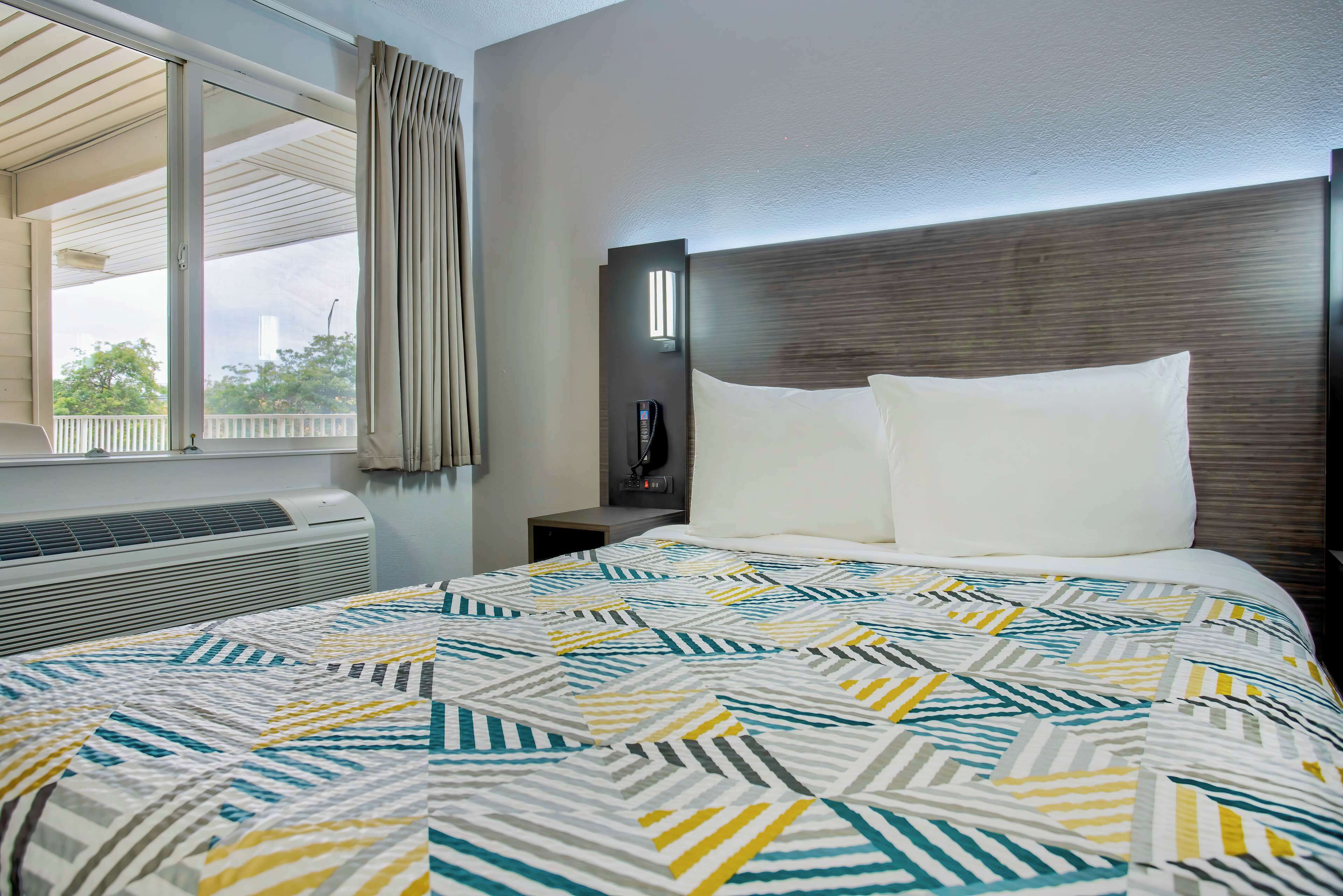 Habitación de hotel con cama, mesita de noche, lámpara y ventana con cortinas.