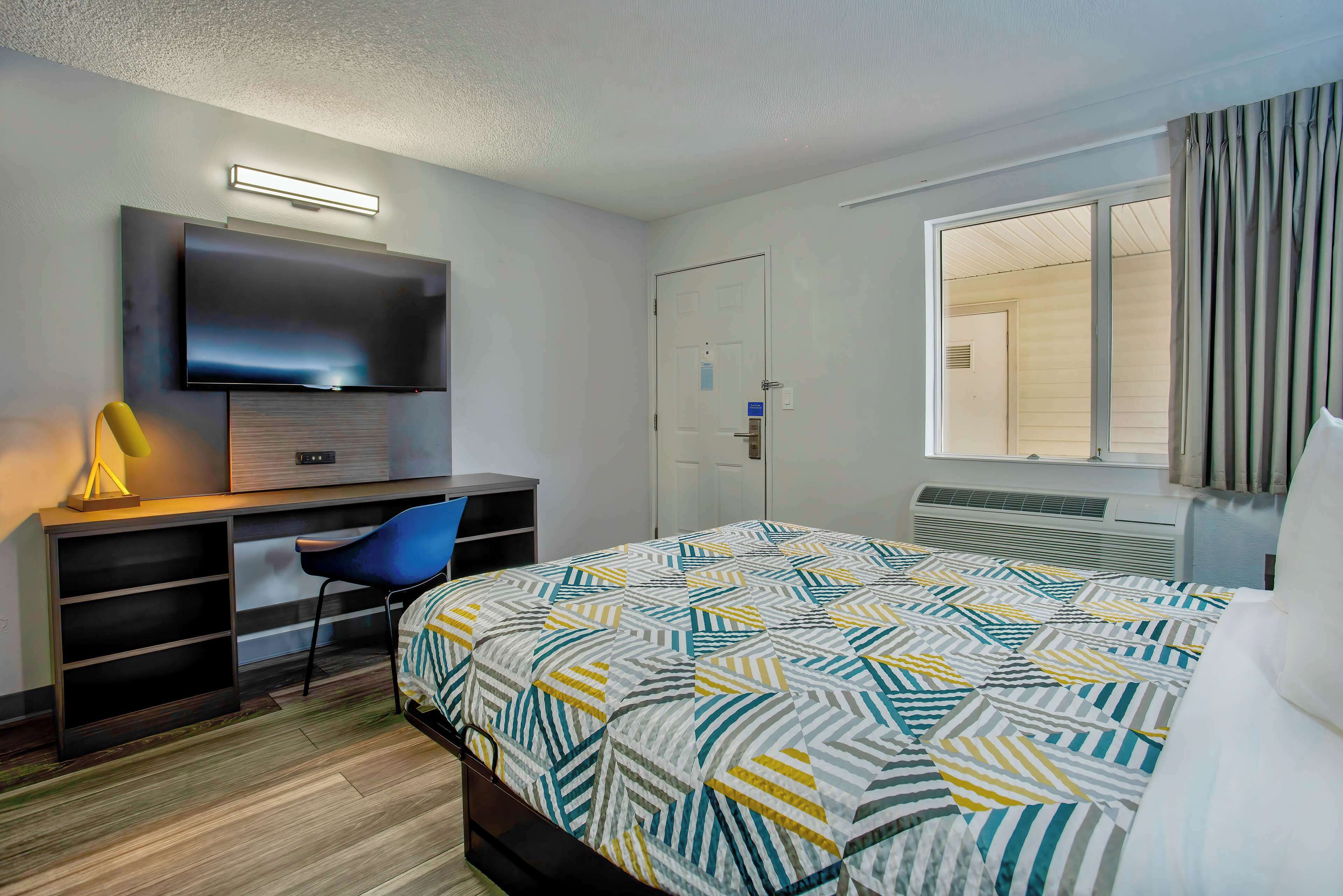 Une chambre d’hôtel comprenant un lit, un bureau avec un téléviseur à écran plat, une chaise et une fenêtre avec des rideaux.
