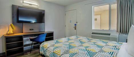 A hotel room with a bed, a desk with a flat-screen TV, a chair, and a window with curtains.