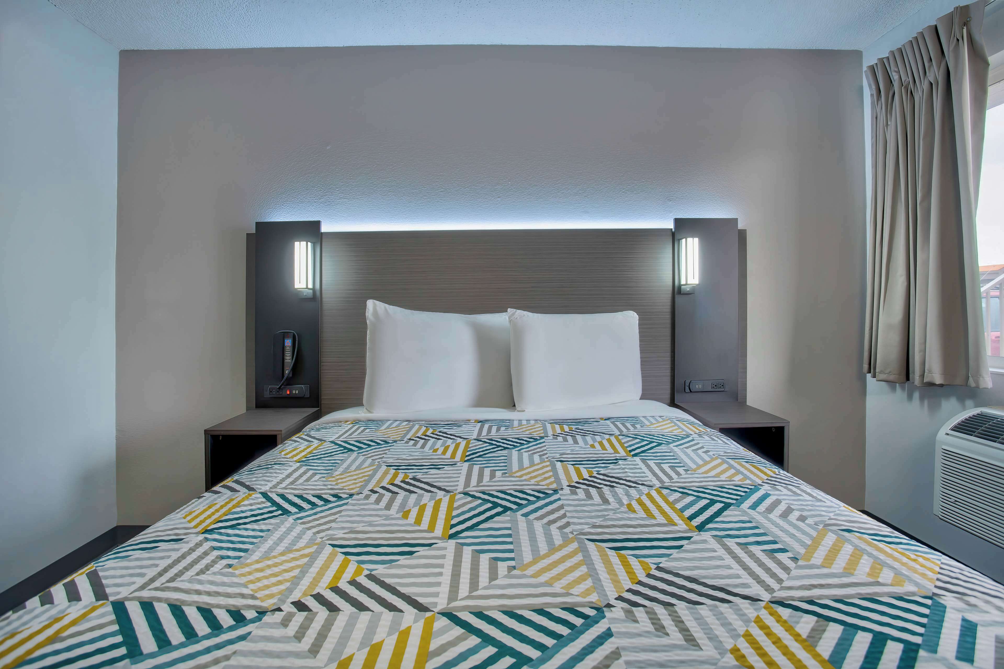 A hotel room with a bed, two bedside tables, a headboard with integrated lighting, a patterned bedspread, and a window with curtains.