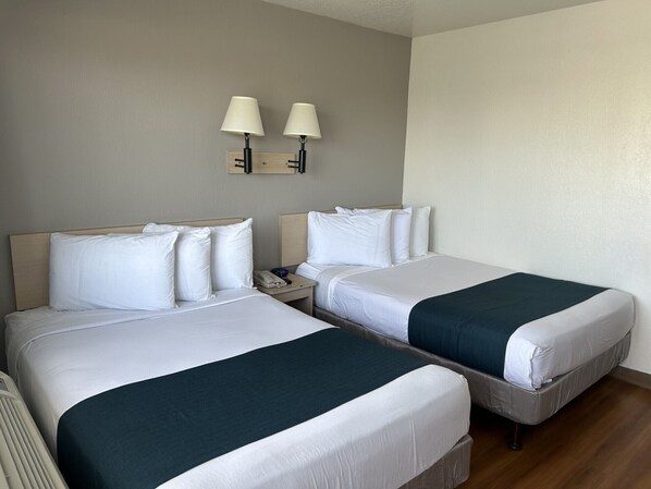 A hotel room with two beds, each with white bedding and a dark green throw. There are two bedside tables with lamps, and a wall-mounted light fixture above the beds.