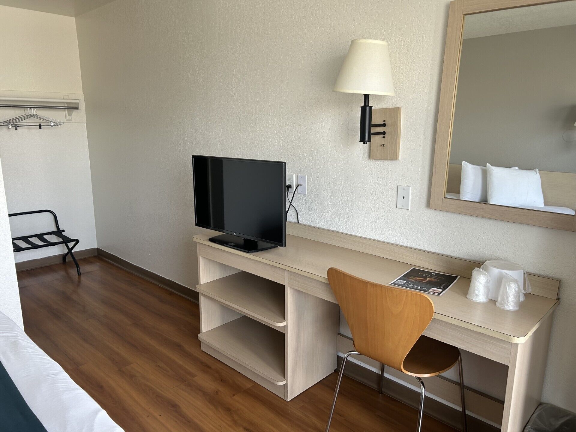 Quarto de hotel com escrivaninha de madeira, televisão, luminária, espelho e uma cadeira.