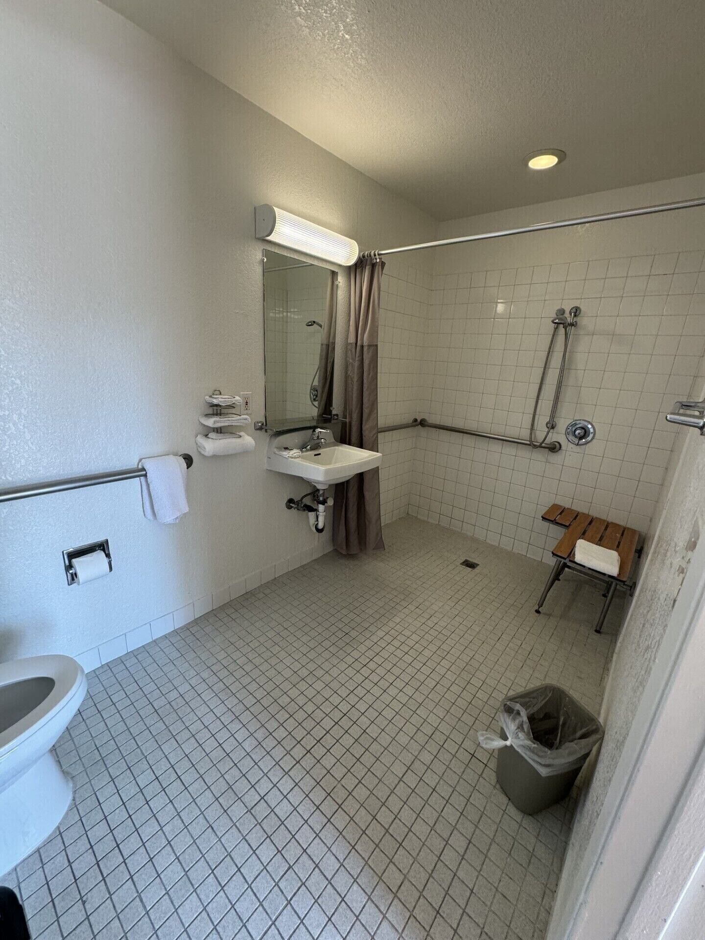 A bathroom with a toilet, sink, mirror, shower, and a bench.