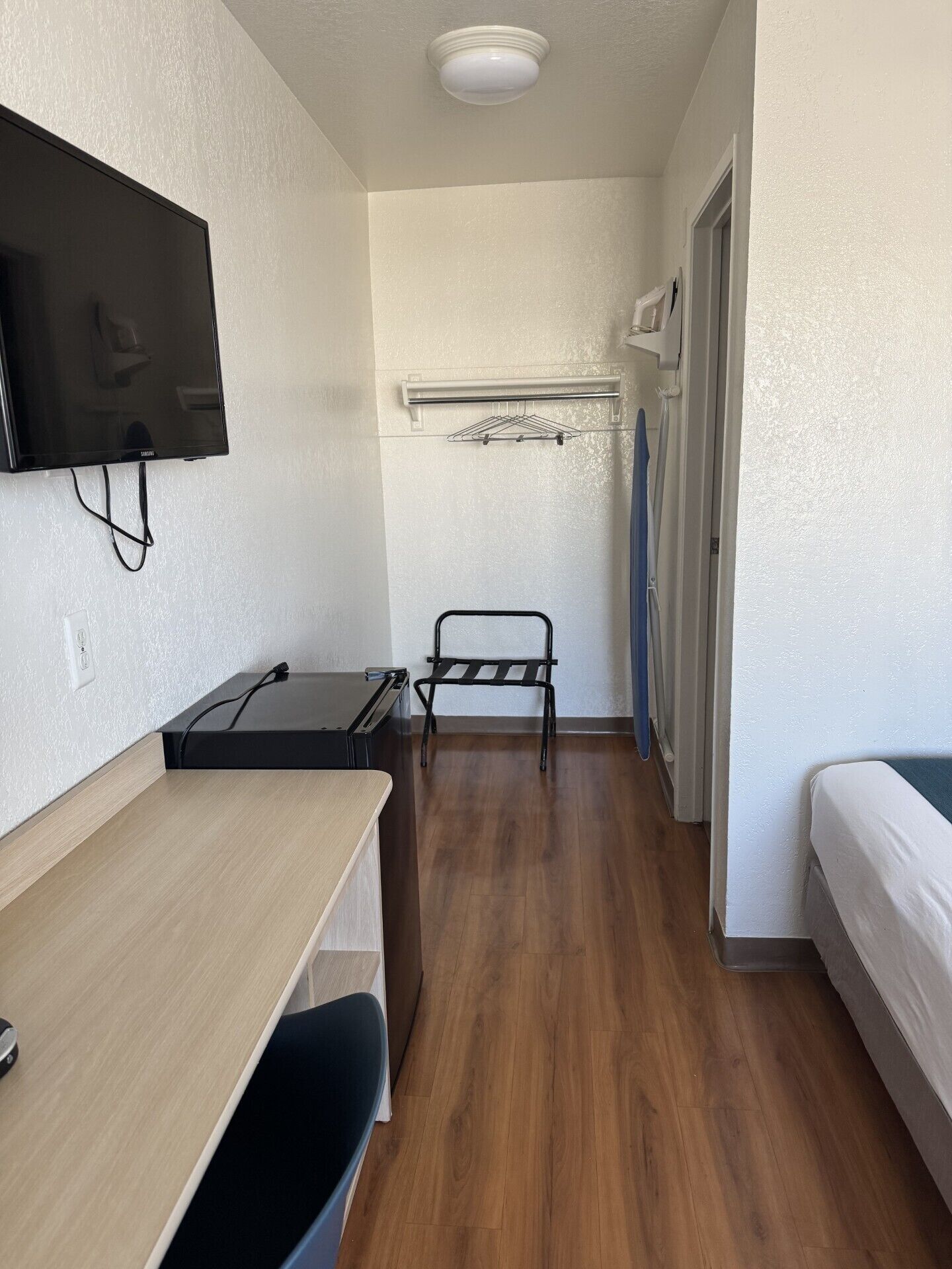 A narrow room with a bed, a desk, a chair, a TV mounted on the wall, and a clothes drying rack.