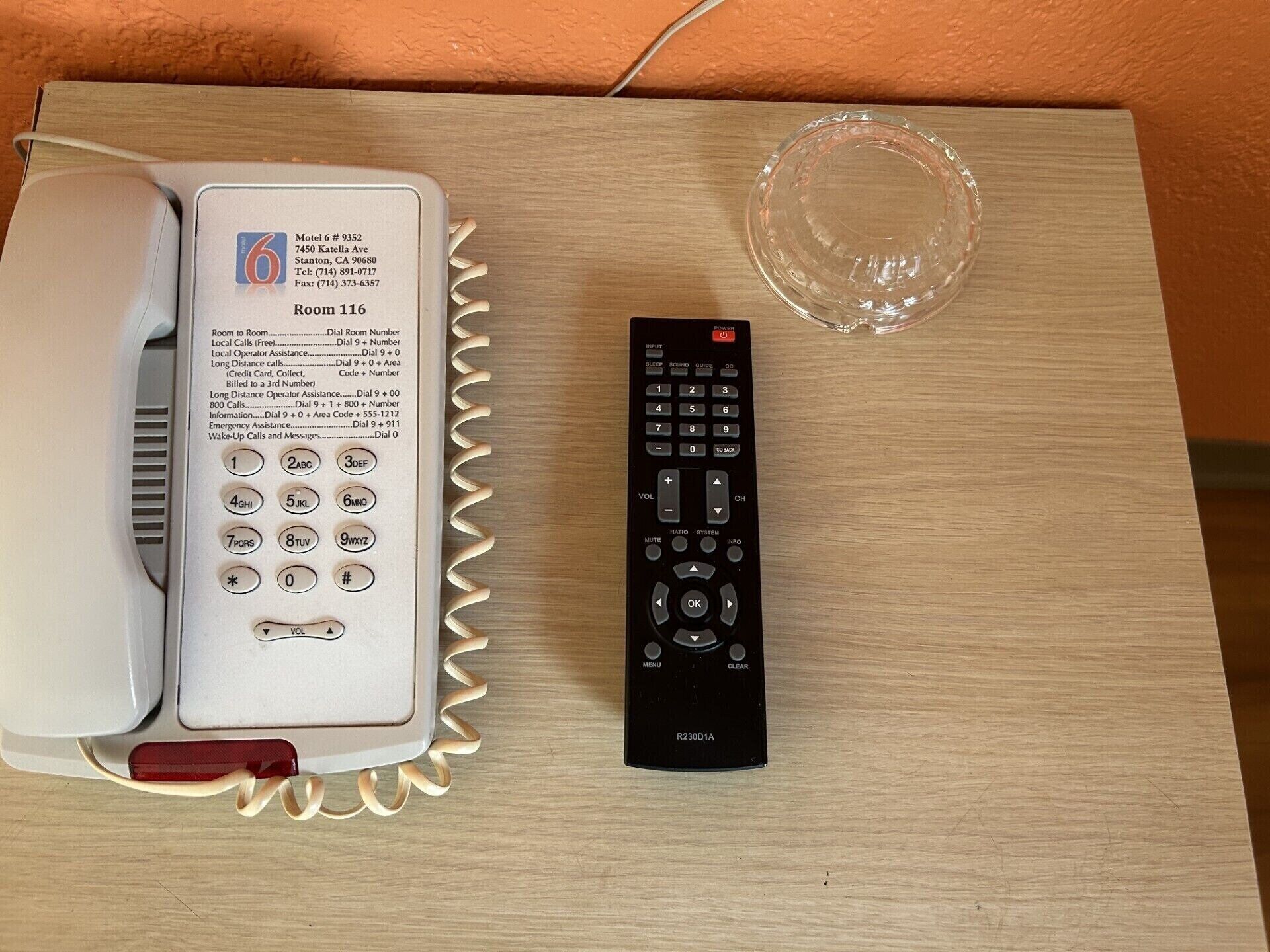 A hotel room with a telephone, a remote control, and a clear glass object on a wooden surface.