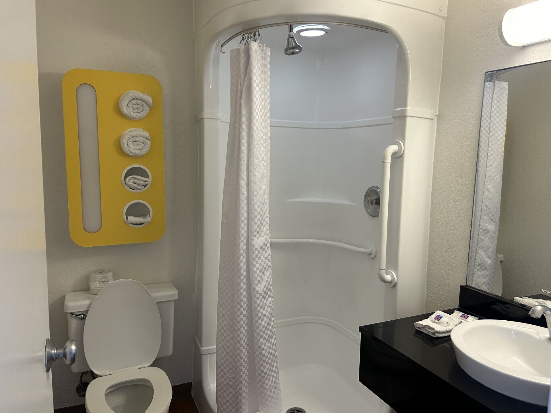 Een badkamer met een wit toilet, een wastafel met een zwart aanrecht, een douche met een geruit gordijn en een gele handdoekhouder met drie handdoeken.